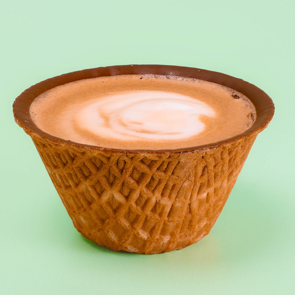 A small textured waffle cone cup filled with a light brown coffee drink topped with a white latte swirl, set against a solid pale green background.