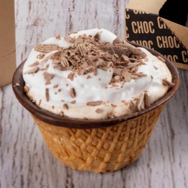 A close-up of a chocolate ice cream cupcake in a waffle cone, topped with white whipped cream and chocolate shavings, with part of its branded box visible in the background.