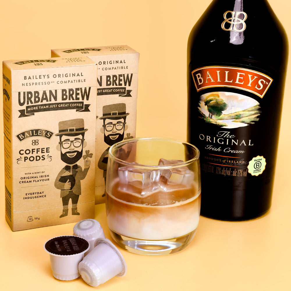 Baileys Urban Brew Nespresso-compatible coffee pods shown with two branded boxes, several loose pods, and a glass of layered coffee with a large bottle of Baileys Irish Cream on a warm beige background.