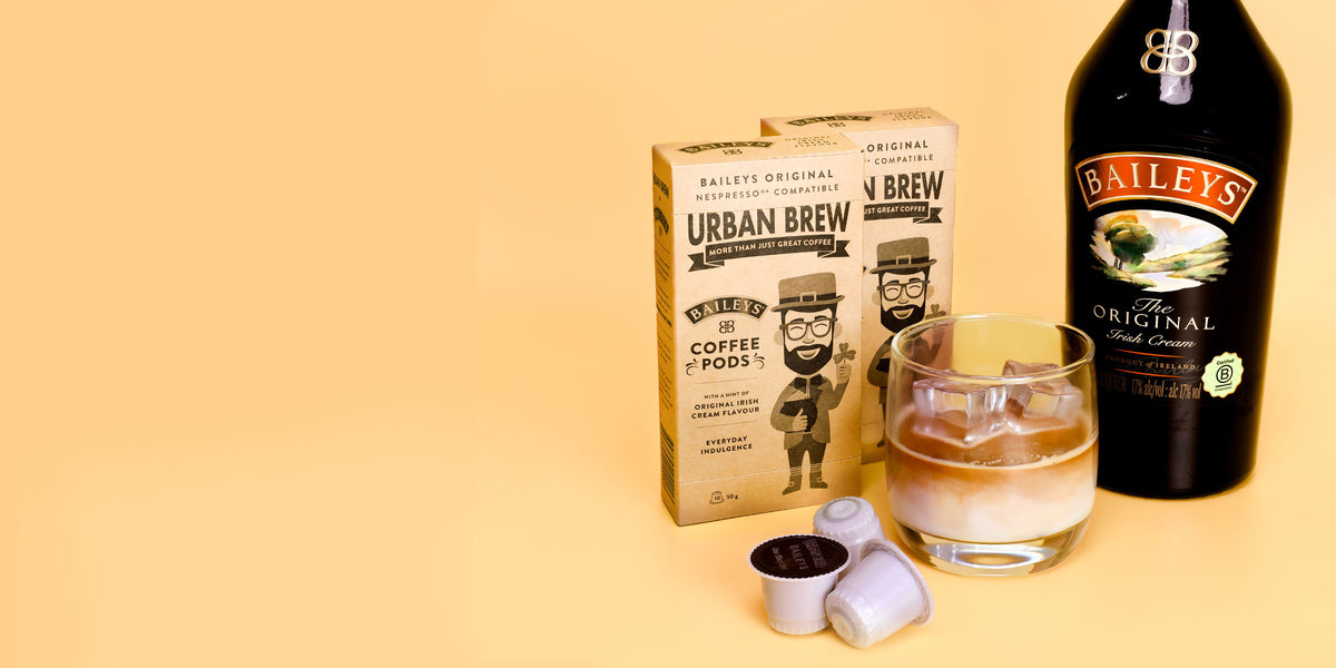 Baileys Urban Brew coffee pods shown with a box of Urban Brew coffee pods, a bottle of Baileys Original Liqueur, a glass of mixed drink, and two coffee pods on a solid beige background.