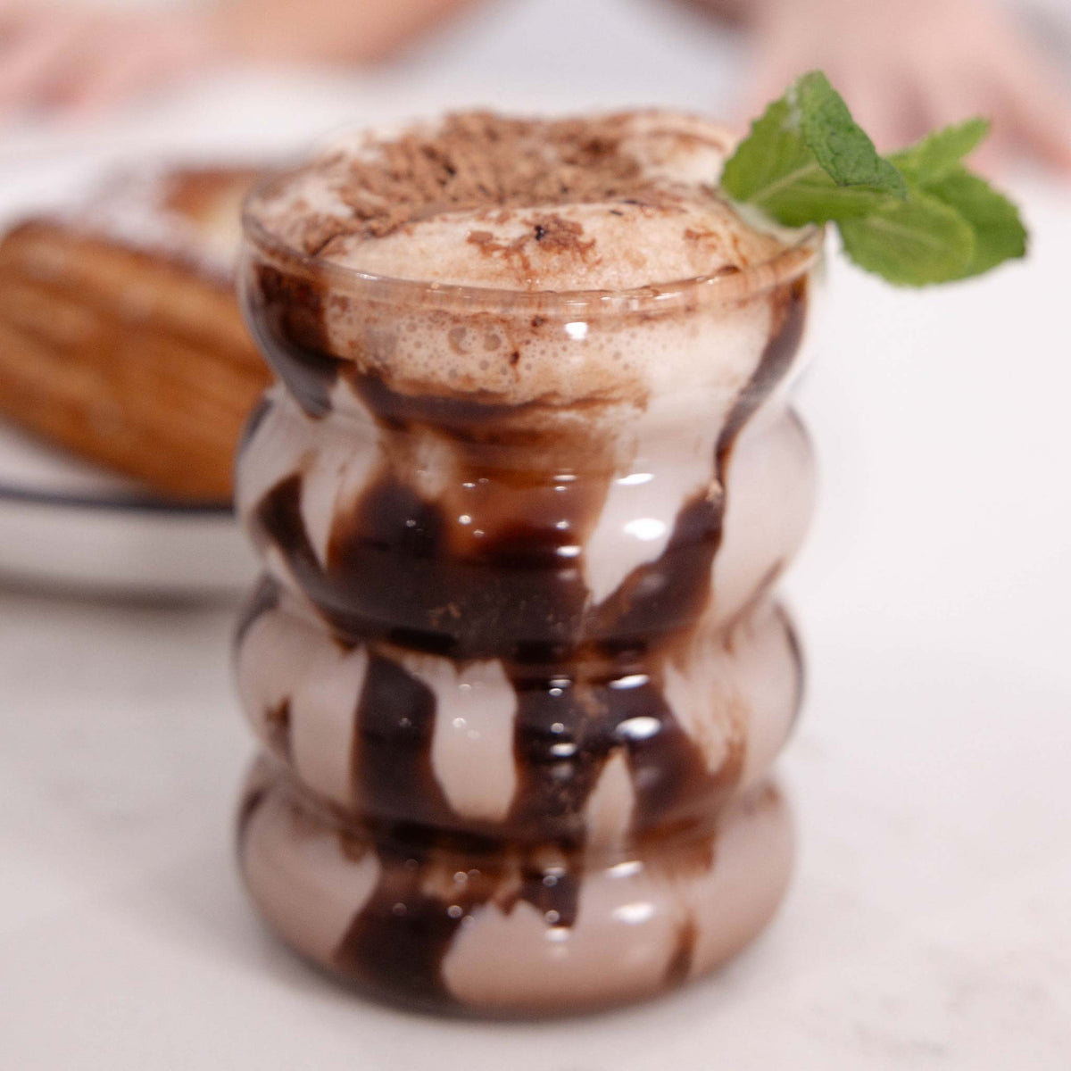 A frothy dessert drink in a stacked, wavy clear glass, topped with whipped cream, cocoa powder, and a mint leaf, with chocolate sauce dripping down the sides. A blurred pastry is visible in the background on a white surface.