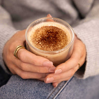 A close-up of a person holding a clear glass cup filled with a light brown latte or coffee drink topped with froth and cocoa powder. The person is wearing a gray knit sweater, blue jeans, and gold rings.