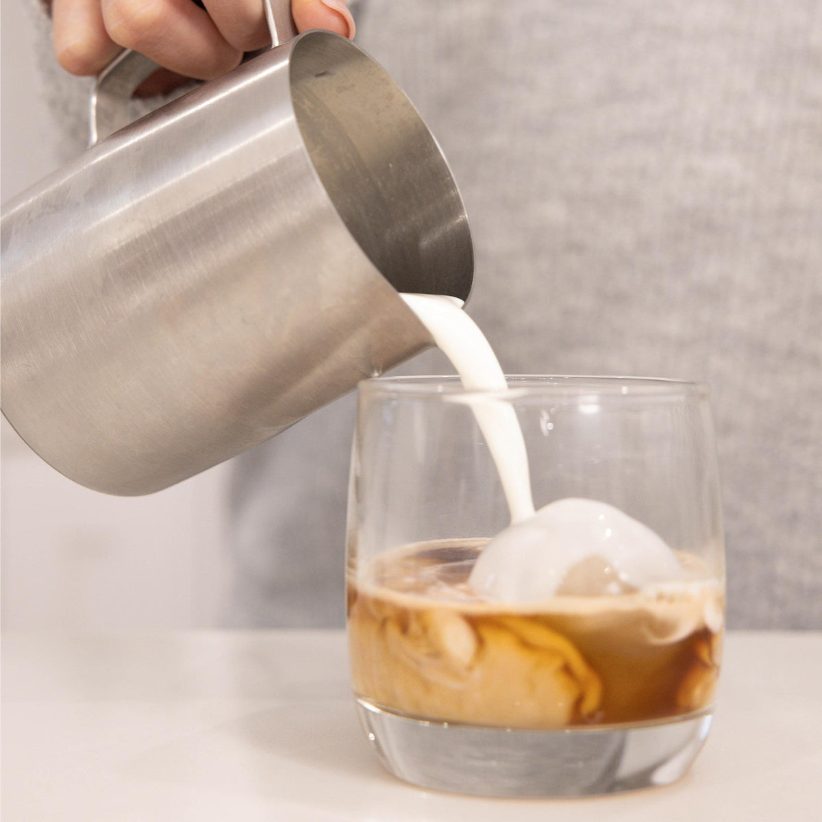 A stainless steel milk pitcher being poured with milk into a clear glass containing an iced coffee drink, shown against a soft gray background.