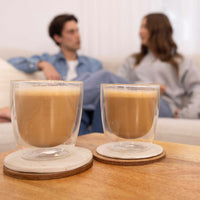 Two clear double-walled glasses filled with a light brown beverage sit on round coasters on a wooden table, with a blurred couple sitting on a sofa in the background.