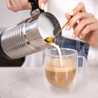 A close-up lifestyle shot of a person pouring steamed milk from a stainless steel pitcher into a double-walled glass mug containing coffee, with a gold spoon resting inside the mug.