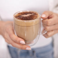 A person holding a clear double-walled glass mug filled with a light brown latte or cappuccino topped with cocoa powder, against a softly blurred white top and blue jeans background.