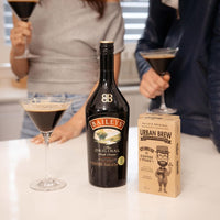 A bottle of Baileys Irish Cream and a box of Urban Brew Coffee Roast sit on a white countertop, with two people in the background holding martini glasses.