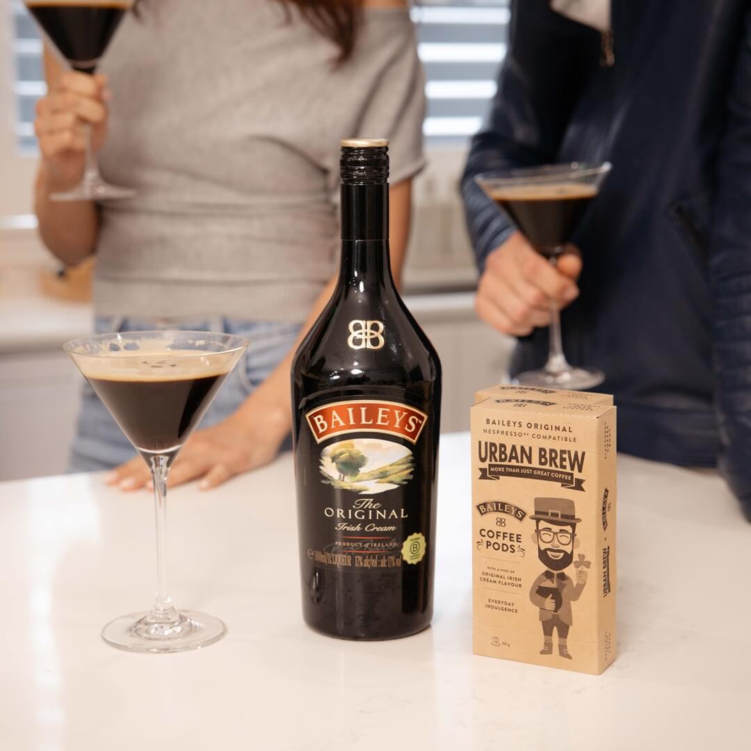A bottle of Baileys Irish Cream and a box of Urban Brew Coffee Roast sit on a white countertop, with two people in the background holding martini glasses.