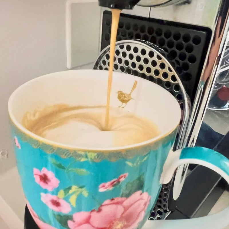 Customer photo: A close-up shot of espresso being poured into a turquoise floral ceramic mug with pink flowers and a gold bird design, set next to a coffee machine.