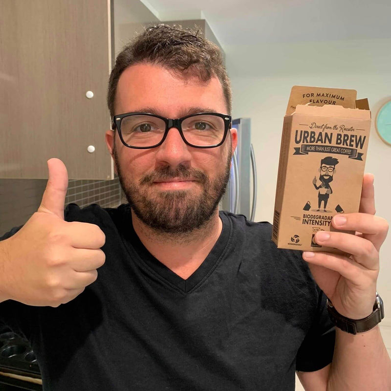 Customer photo: A man wearing black-rimmed glasses and a black t-shirt gives a thumbs-up while holding up a box of Urban Brew Roastware Intensity coffee in a kitchen setting.