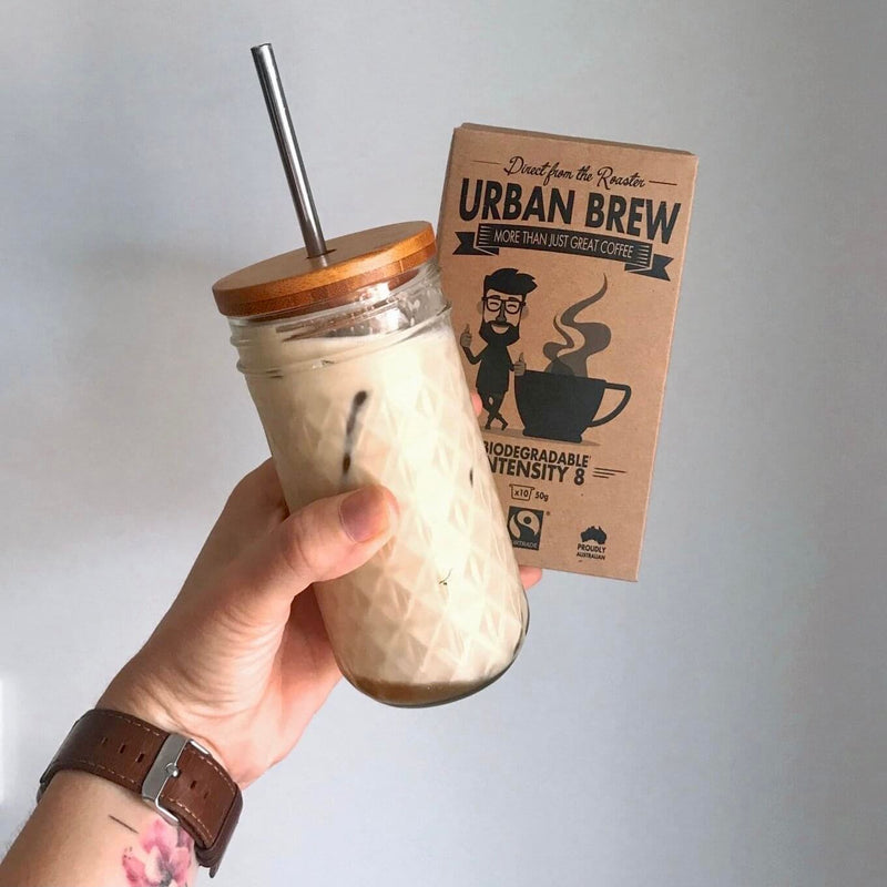 A hand holds a glass jar mug filled with iced coffee, featuring a bamboo lid and metal straw, next to its brown cardboard packaging box labeled 'Urban Brew'.