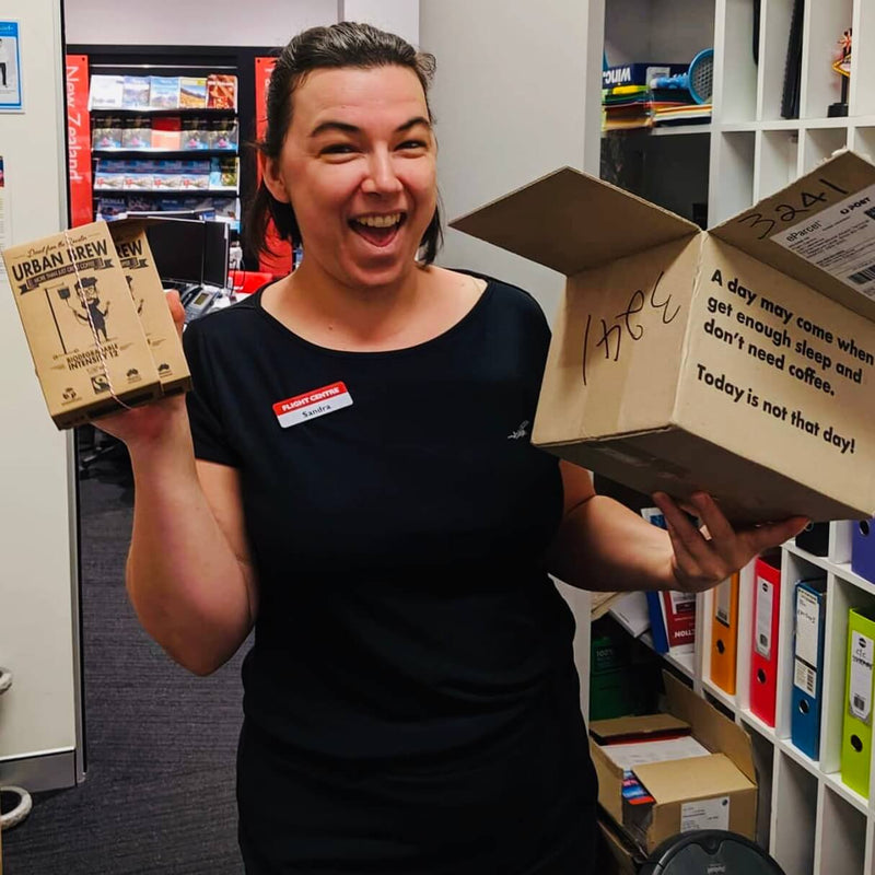 Customer photo: A smiling woman in a black t-shirt holding two cardboard boxes in an office setting. One box is labeled 'URBAN BREW' and the other has handwritten text on it.