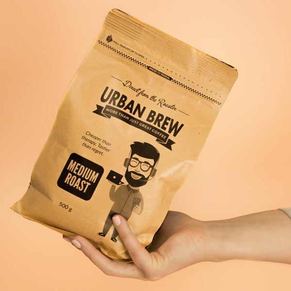 A hand holding a 500g bag of Urban Brew Medium Roast coffee against a solid peach background.