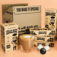 Urban Brew coffee capsule packaging arranged with a cardboard shipping box, multiple paperboard product boxes, two single-serve capsules, and a double-walled glass cup filled with latte art coffee, set against a peach studio background.