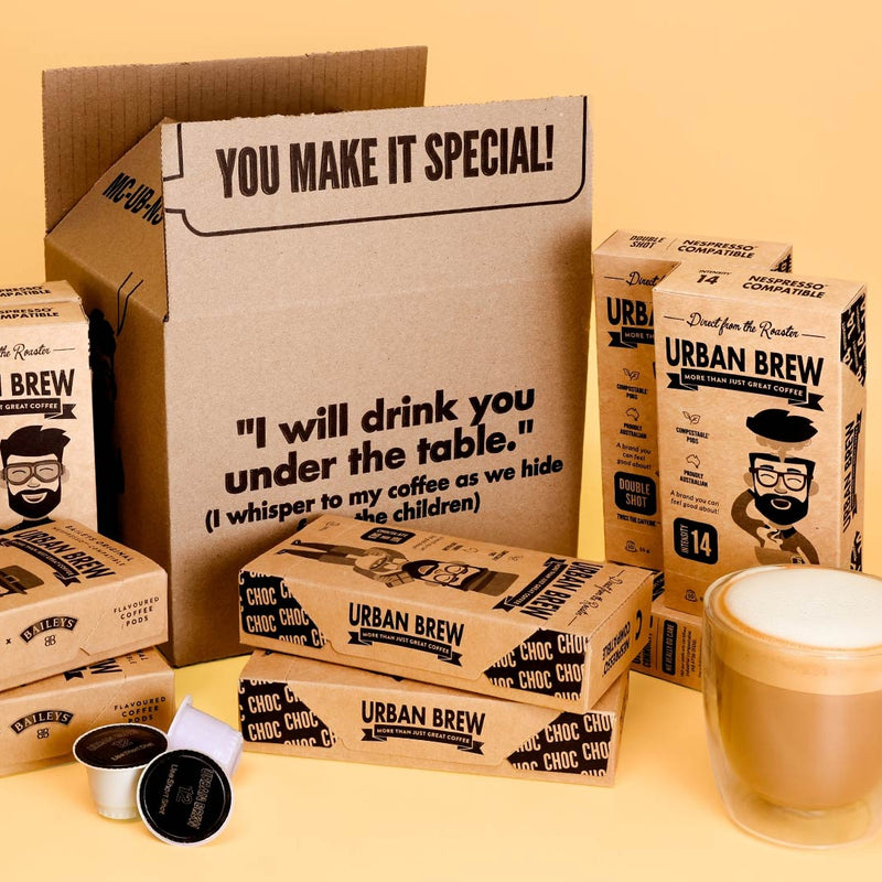 Urban Brew coffee capsule packaging arranged around an open cardboard box on a warm beige background, with a glass of latte and two coffee capsules in the foreground.