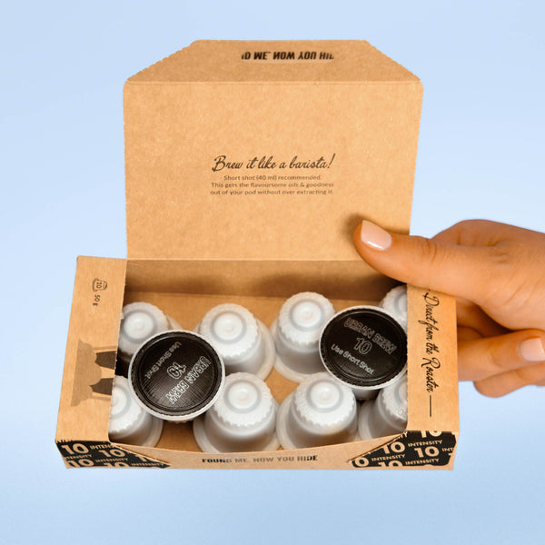 A hand holds open a small kraft paper box containing multiple single-serve coffee pods. The box lid is printed with coffee-themed text, and the pods inside are visible through a clear window.