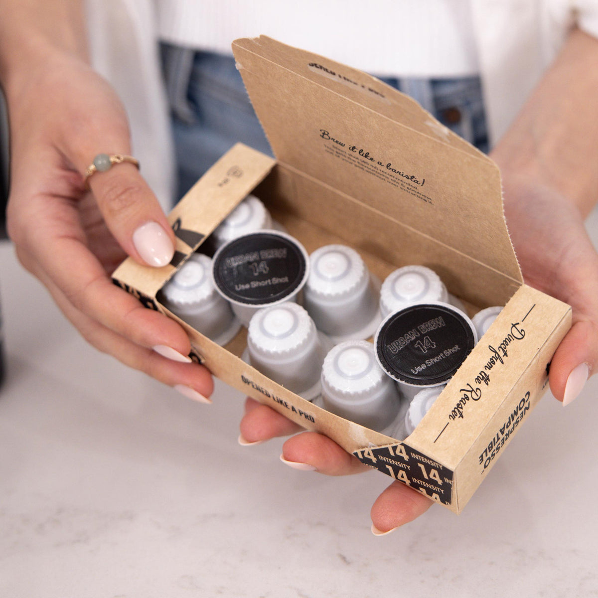 A person holds an open kraft paper box containing multiple small coffee pods. The box is branded 'Coffee at the Table' and 'Café Artisanal', with two pods showing black lids labeled 'Line Stout (Coffee)'.